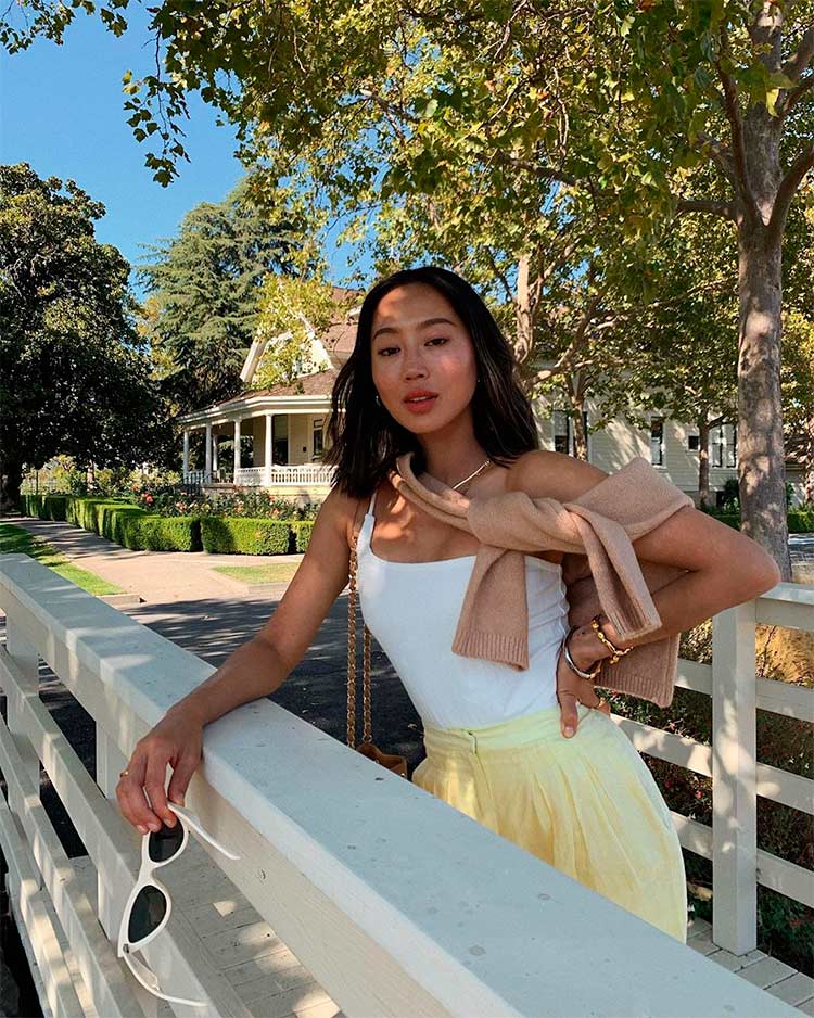 Aimee Song, una de las influencers de Instagram que más cobra, posando al lado de una casa de campo, vestida con un top blanco, unos pantalones amarillos de tiro alto y un jersey color salmón anudado al hombro. 